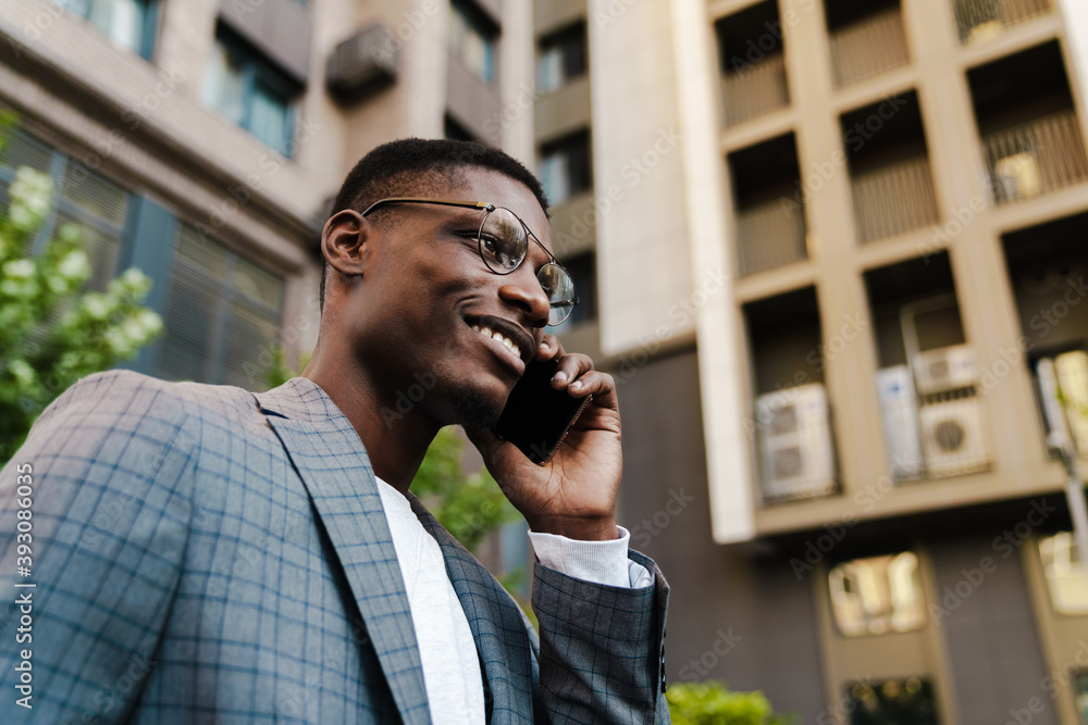 Joyful african american man talking on mobile phone while walking on street - obrazy, fototapety, plakaty 