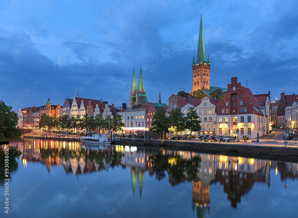 Fototapeta premium Lubeck, Germany. Evening of the Lubeck Old Town with two towers of St. Mary's Church and single tower of St. Peter's Church. View from the shore of Trave river.