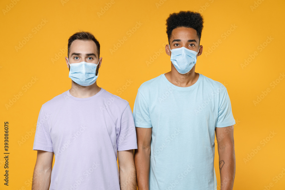 Young friends european african american men 20s in purple blue casual t-shirts sterile face mask to safe from coronavirus covid-19 looking camera isolated on yellow colour background studio portrait.