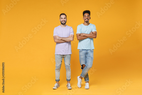 Full length of smiling young two friends european african american men 20s in purple blue casual t-shirts holding hands crossed looking camera isolated on yellow colour background studio portrait.