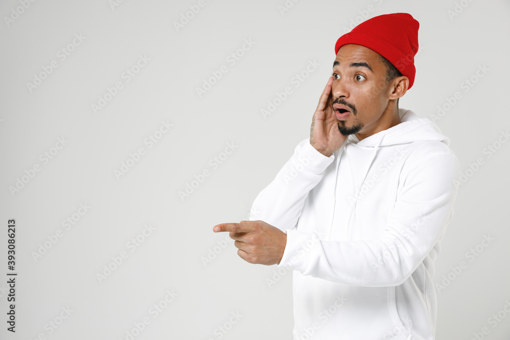 Shocked young african american man 20s wearing basic casual streetwear hoodie put hand on cheek pointing index finger aside on mock up copy space isolated on white colour background, studio portrait. Shocked young african american man 20s wearing basic casual streetwear hoodie put hand on cheek pointing index finger aside on mock up copy space isolated on white colour background, studio portrait.