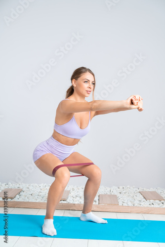 Attractive strong fit blonde in pink sportswear doing squats with elastic band in cozy interior.