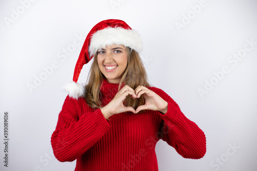 Young pretty blonde woman wearing a red casual sweater and a christmas hat over white background smiling in love showing heart symbol and shape with hands