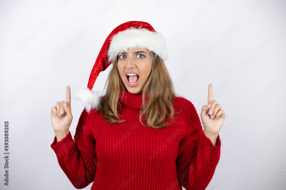 Young pretty blonde woman wearing a red casual sweater and a christmas hat over white background amazed and surprised looking at the camera and pointing up with fingers and raised arms Young pretty blonde woman wearing a red casual sweater and a christmas hat over white background amazed and surprised looking at the camera and pointing up with fingers and raised arms