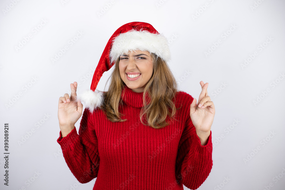 Young pretty blonde woman wearing a red casual sweater and a christmas hat over white background gesturing finger crossed smiling with hope and looking side Young pretty blonde woman wearing a red casual sweater and a christmas hat over white background gesturing finger crossed smiling with hope and looking side