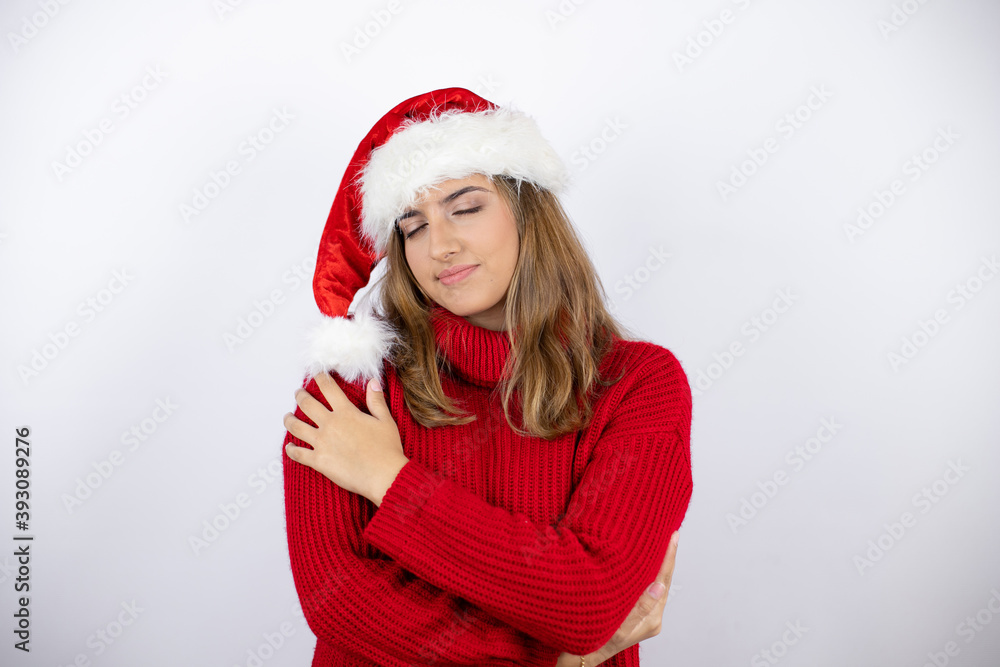Young pretty blonde woman wearing a red casual sweater and a christmas hat over white background hugging oneself happy and positive, smiling confident Young pretty blonde woman wearing a red casual sweater and a christmas hat over white background hugging oneself happy and positive, smiling confident