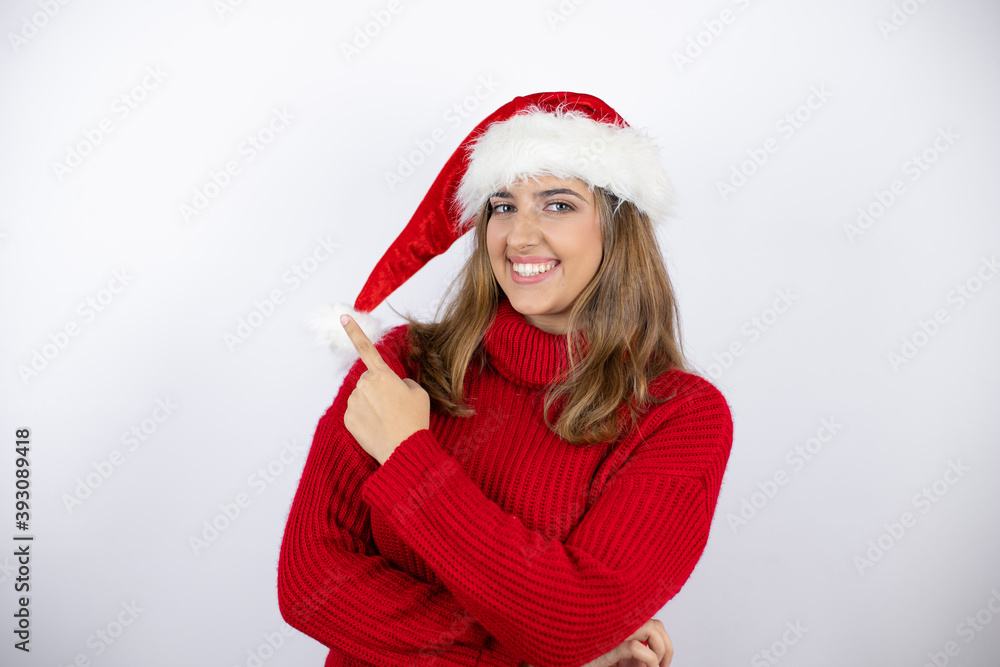 Young pretty blonde woman wearing a red casual sweater and a christmas hat over white background smiling and pointing with hand and finger to the side Young pretty blonde woman wearing a red casual sweater and a christmas hat over white background smiling and pointing with hand and finger to the side