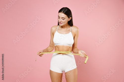 Smiling pretty attractive thin sports young brunette woman 20s in white underwear standing posing measuring waist with measure tape isolated on pastel pink colour wall background, studio portrait. © ViDi Studio