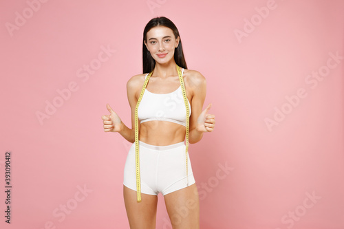 Smiling young brunette woman 20s in white underwear with strong fit body posing hold measure tape showing thumbs up looking camera isolated on pastel pink colour wall background, studio portrait.