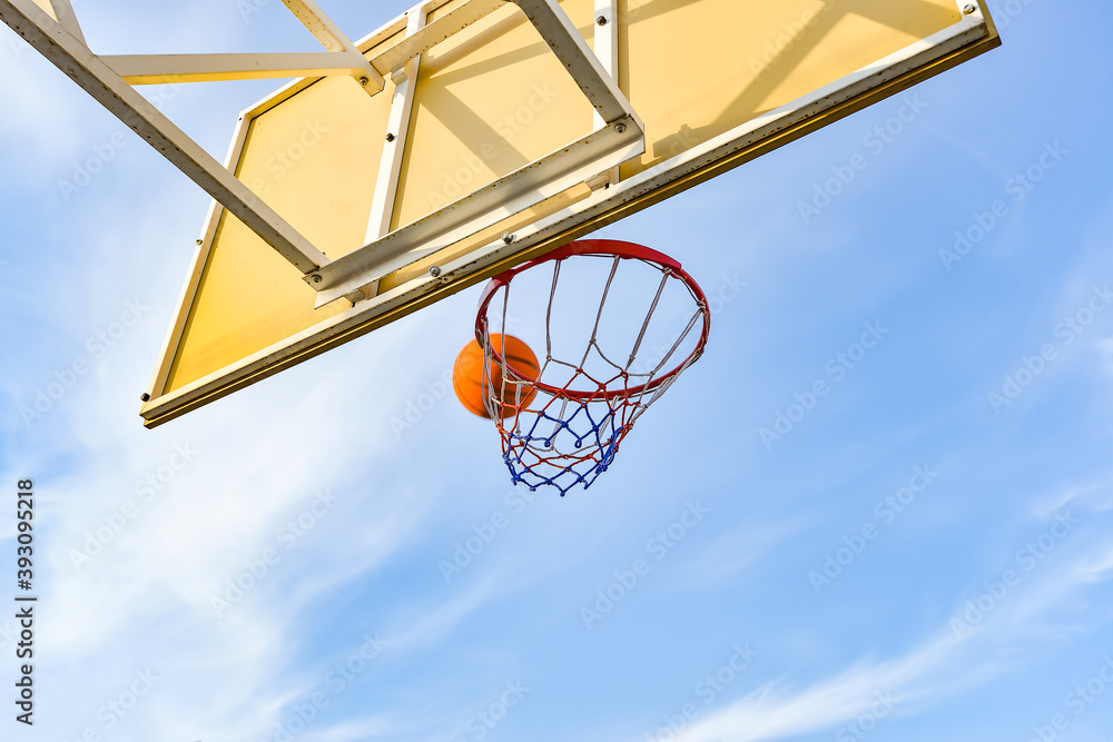 A basketball ball flies into a hoop with a net against a blue cloudy sky. Sports activities on the playground. A basketball ball flies into a hoop with a net against a blue cloudy sky. Sports activities on the playground.