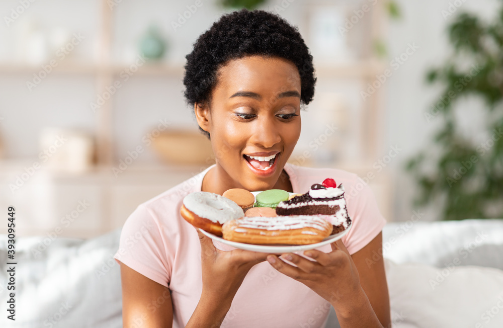 Diet breakdown and unhealthy nutrition concept. Young African American lady holding plate of sweet pastries at home Diet breakdown and unhealthy nutrition concept. Young African American lady holding plate of sweet pastries at home