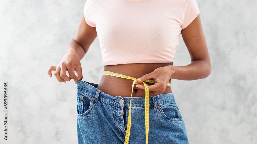 Black woman in old big jeans measuring her waist, showing results of slimming diet or liposuction, grey background - obrazy, fototapety, plakaty 