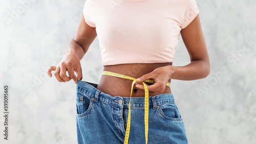 Black woman in old big jeans measuring her waist, showing results of slimming diet or liposuction, grey background