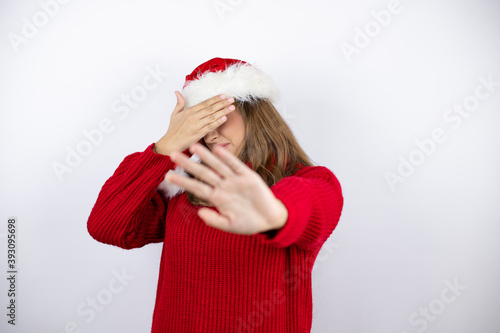 Young pretty blonde woman wearing a red casual sweater and a christmas hat over white background covering eyes with hands and doing stop gesture with sad and fear expression.