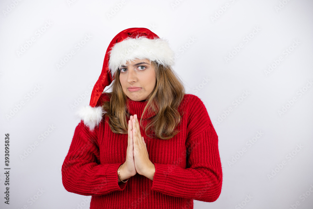 Young pretty blonde woman wearing a red casual sweater and a christmas hat over white background begging and praying with hands together with hope expression on face very emotional and worried Young pretty blonde woman wearing a red casual sweater and a christmas hat over white background begging and praying with hands together with hope expression on face very emotional and worried
