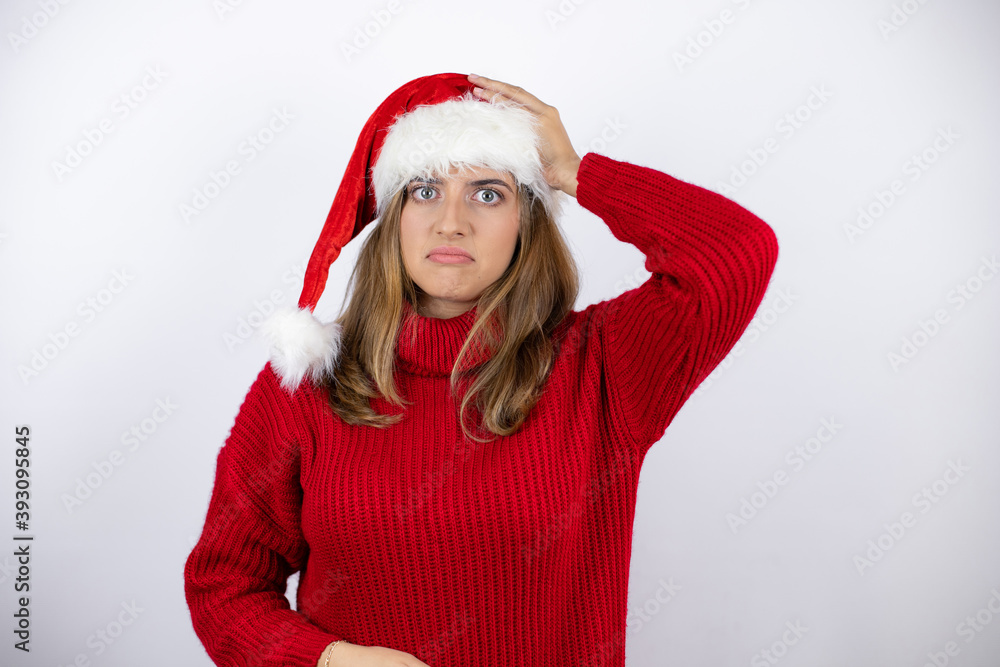 Young pretty blonde woman wearing a red casual sweater and a christmas hat over white background putting one hand on her head smiling like she had forgotten something Young pretty blonde woman wearing a red casual sweater and a christmas hat over white background putting one hand on her head smiling like she had forgotten something