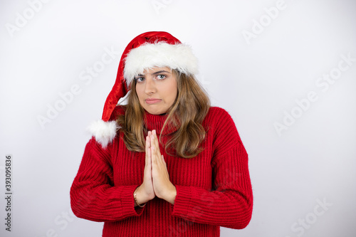 Young pretty blonde woman wearing a red casual sweater and a christmas hat over white background begging and praying with hands together with hope expression on face very emotional and worried