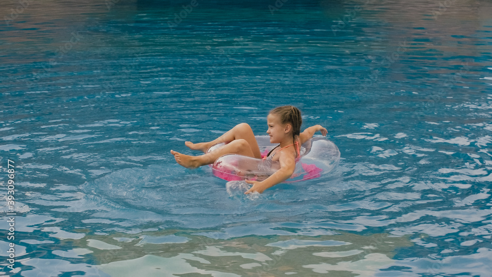 The little cute girl have fun in the pool. The child enjoy summer vacation in a swimming pool jumping, spinning, splash water. Happy childhood.