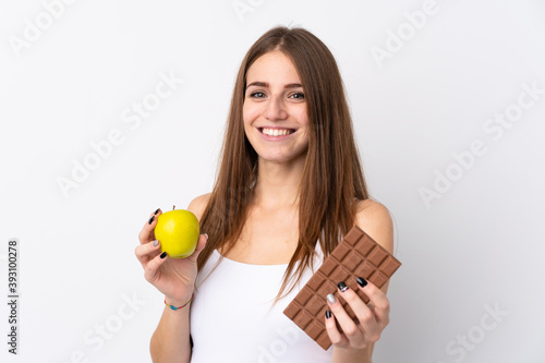 Young woman over isolated white background taking a chocolate tablet in one hand and an apple in the other
