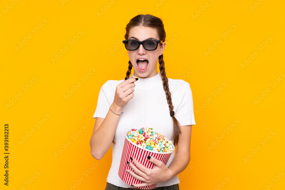 Obraz premium Young woman over isolated yellow background with 3d glasses and holding a big bucket of popcorns