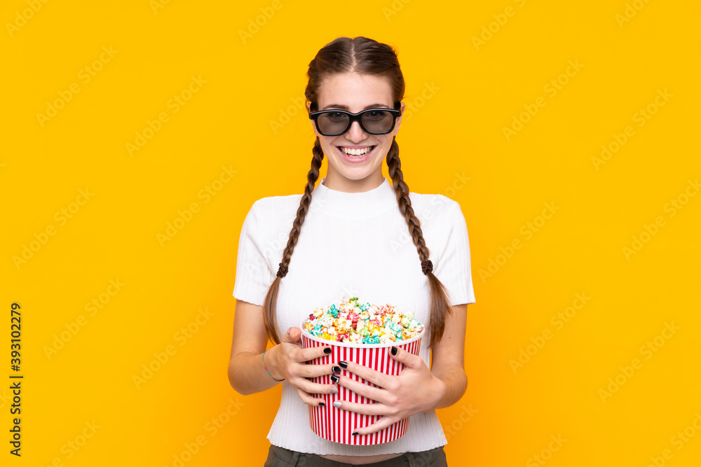 Young woman over isolated yellow background with 3d glasses and holding a big bucket of popcorns