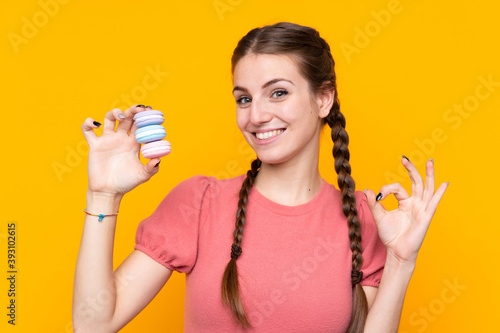 Young woman over isolated yellow background holding colorful French macarons and showing ok sign with fingers