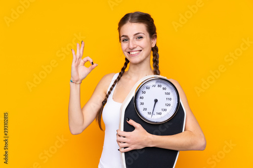Young woman over isolated yellow background holding a weighing machine and doing OK sign