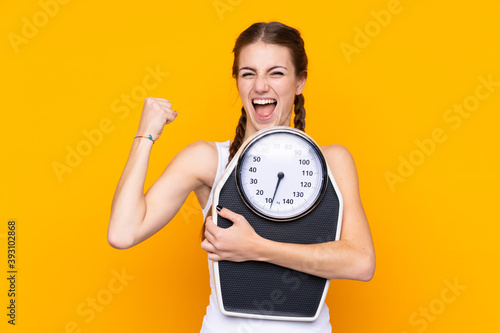 Young woman over isolated yellow background with weighing machine and doing victory gesture