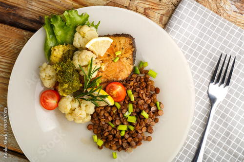 Baked fish with broccoli and lentils in a white plate with rosemary and lemon on a napkin next to a fork.
