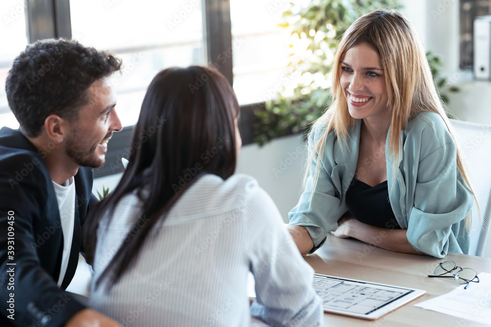 Beautiful real-estate agent showing house plans on electronic tablet while talking to the couple about buying the house in the office. Beautiful real-estate agent showing house plans on electronic tablet while talking to the couple about buying the house in the office.