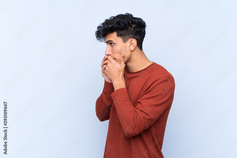 Young Argentinian man over isolated blue background covering mouth and looking to the side Young Argentinian man over isolated blue background covering mouth and looking to the side