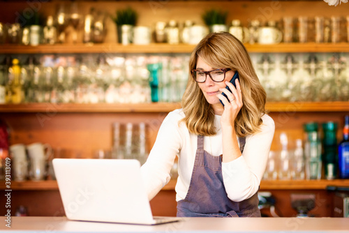 Shot of smiling female waitress using mobile phone and laptop while standing at the counter and working in the cafe. Confident mature cafe owner businesswoman standing at cafeteria