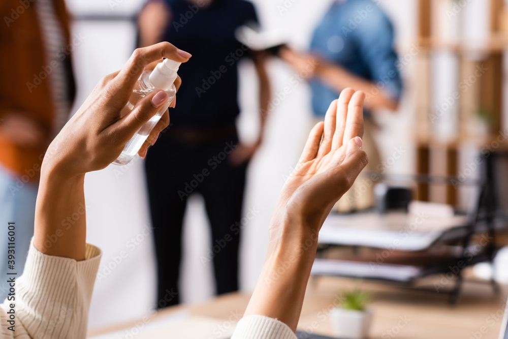 Obraz premium cropped view of african american woman spraying sanitizer on hands near colleagues talking on blurred background