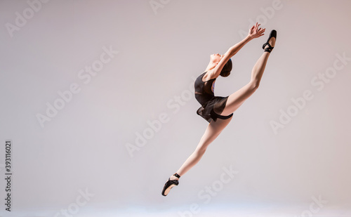 A young ballerina in a black swimsuit and pointes jumps against a bright background.