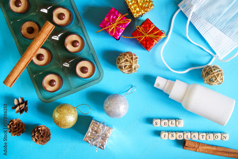 Christmas toys, gifts and chocolates lie on a blue background next to a protective medical mask and sanitizer. New Year's self-isolation.