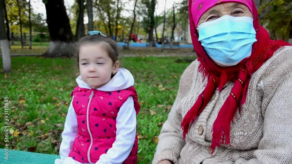 An old grandmother is resting with a child on a bench in the park. A grandmother and a child wearing masks at a social distance during the coronavirus. Walking child with grandmother in autumn An old grandmother is resting with a child on a bench in the park. A grandmother and a child wearing masks at a social distance during the coronavirus. Walking child with grandmother in autumn