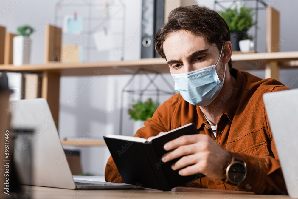 young businessman in medical mask looking in notebook at workplace on blurred background