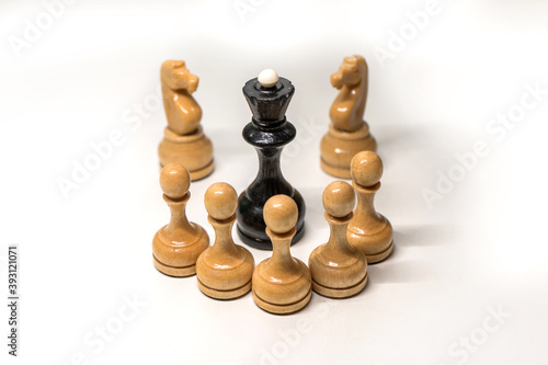 Wooden chess pieces. The black Queen is surrounded by white pawns and two knights looking at the center, focus on the black Queen, on a white background