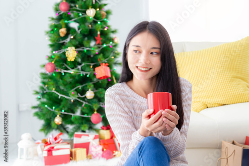 Happy asian woman holding red coffee cup on hands with Christmas tree background. Merry Christmas and New Year concept.