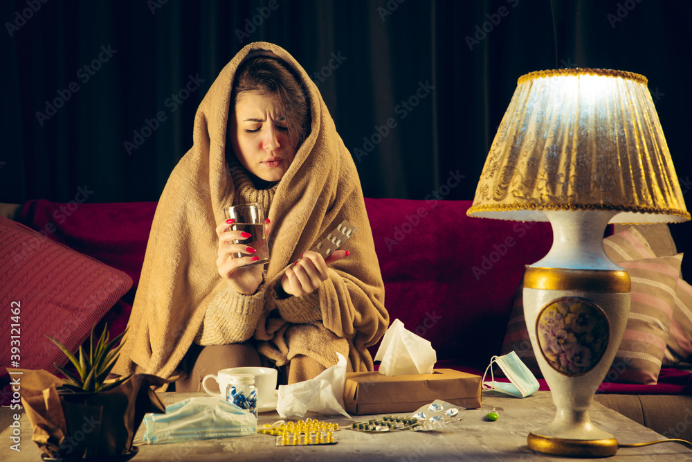 Taking pills. Young woman wrapped in a plaid looks sick, ill, sneezing and coughing sitting on sofa at home indoors. Healthcare and medicine, ill prevention, seasonal illness symptoms and self Taking pills. Young woman wrapped in a plaid looks sick, ill, sneezing and coughing sitting on sofa at home indoors. Healthcare and medicine, ill prevention, seasonal illness symptoms and self