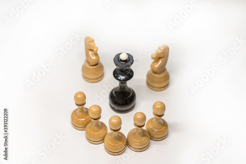 Wooden chess pieces. The black Queen is surrounded by white pawns and two knights looking at the center, focus on the black Queen on top of her head, on a white background. The camera looks from above