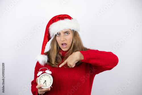 Young pretty blonde woman wearing a red casual sweater and a christmas hat over white background holding and pointing a clock