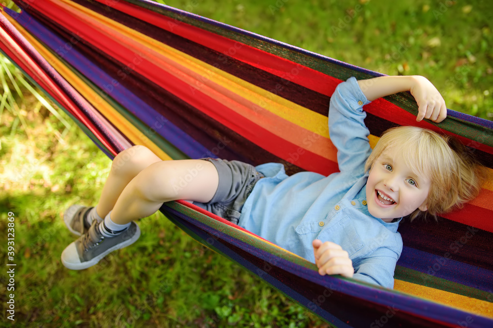 Cute little blond caucasian boy relaxing and having fun in multicolored hammock in backyard or outdoor playground. Summer active leisure for kids. Child swinging on hammock. Cute little blond caucasian boy relaxing and having fun in multicolored hammock in backyard or outdoor playground. Summer active leisure for kids. Child swinging on hammock.