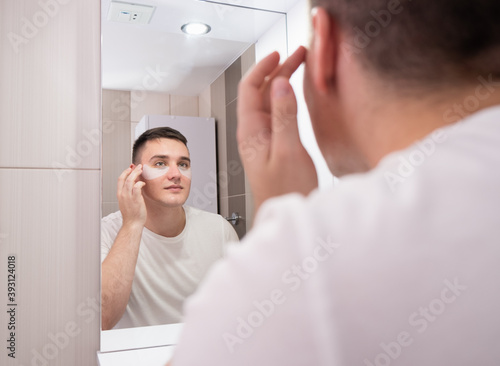Fototapeta Naklejka Na Ścianę i Meble -  Man look in bathroom mirror use under eye anti-wrinkle patches, millennial male do morning facial procedures with hydrogel beauty product, skincare concept
