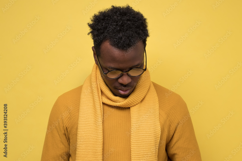 Fototapeta premium Young distressed african man wearing glasses looking down. Studio shot on yellow wall.