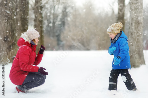 Little boy and mother having fun playing with fresh snow. Snow fight. People dressed in warm clothes  hat  hand gloves and scarf. Active outdoors leisure for family with child in snowy winter day.