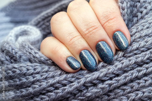 Female hand with gray knitted scarf with beautiful manicure - blue glittered nails. Selective focus. Closeup view