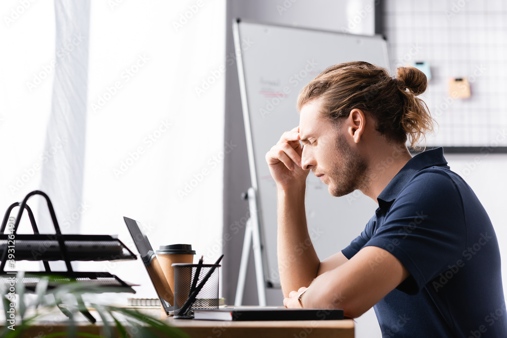 Thoughtful office worker with closed eyes sitting at workplace table near laptop with blurred plant on background Thoughtful office worker with closed eyes sitting at workplace table near laptop with blurred plant on background