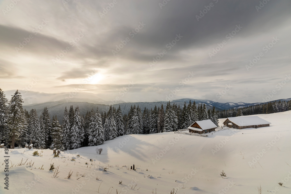 Winter mountain landscape. Old wooden houses on snowy clearing on background of mountain ridge, spruce forest and cloudy sky. Happy New Year and Merry Christmas card. Winter mountain landscape. Old wooden houses on snowy clearing on background of mountain ridge, spruce forest and cloudy sky. Happy New Year and Merry Christmas card.