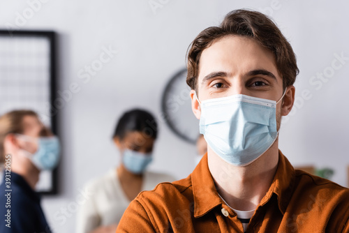 young businessman in protective mask looking at camera while multiethnic colleagues standing on blurred background